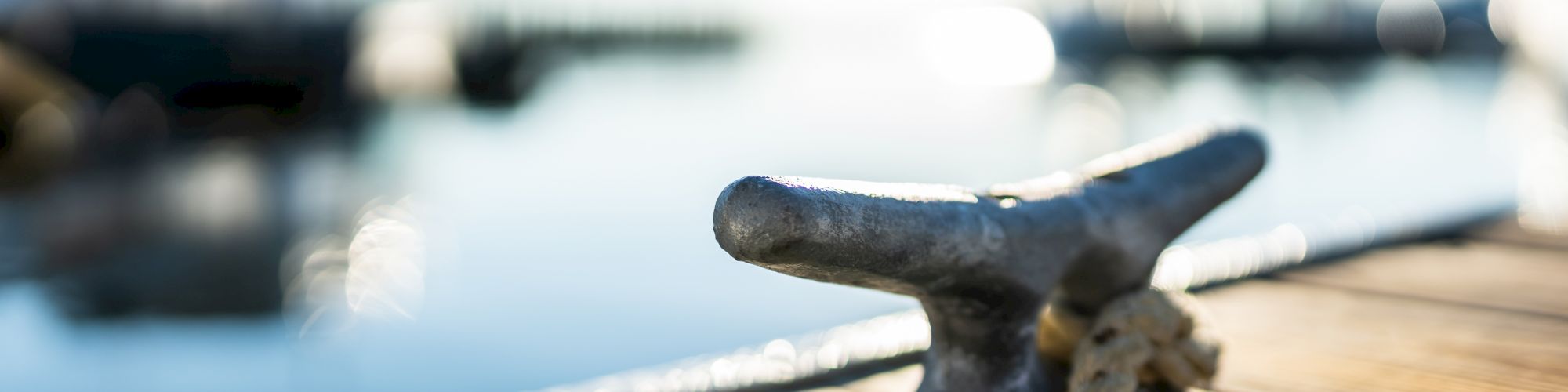 A metal cleat with a tied rope sits on a wooden dock by the water, with blurred boats and a sunny sky in the background.